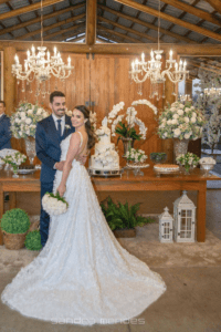 O vestido de noiva reflete sua personalidade: delicada e romântica. Com alças finas e um decote V profundo, o grande destaque do modelo são as aplicações de flores delicadas, que criam um efeito quase de um jardim encantado, onde cada flor parece brotar suavemente, refletindo a beleza e a leveza do amor que ela e Alexandre compartilham.